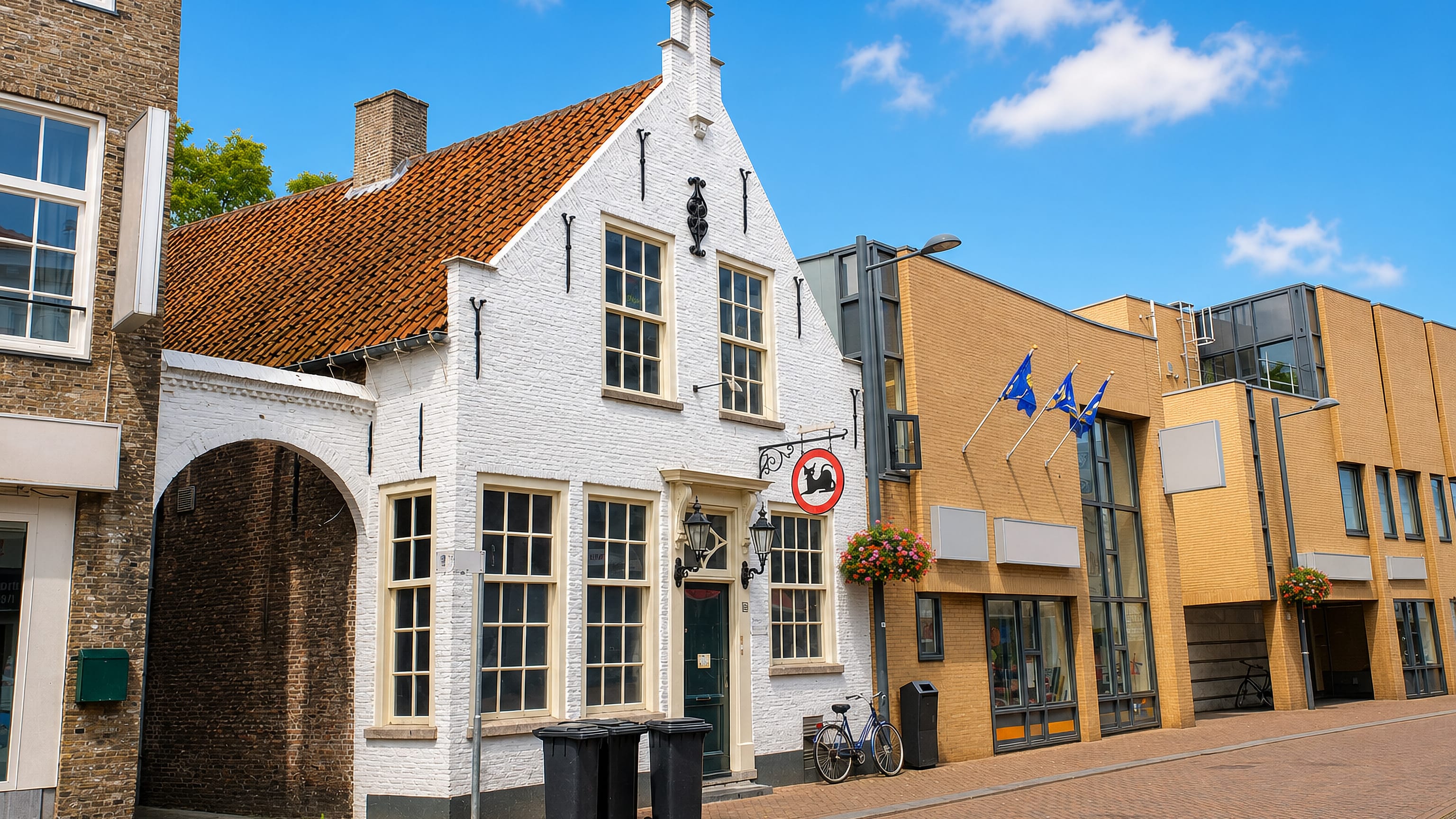 White stepped-gable house on a brick-paved street in Roosendaal, Netherlands, beside modern shopfronts.