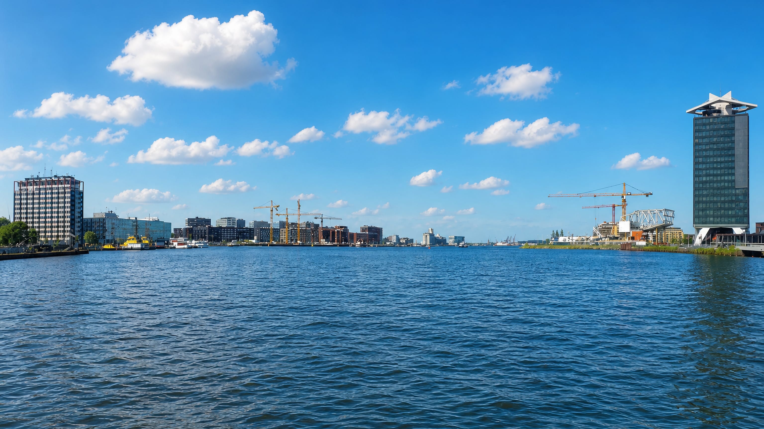 Amsterdam's IJ waterfront under a wide blue sky, with a star-topped riverside tower and dockside cranes.