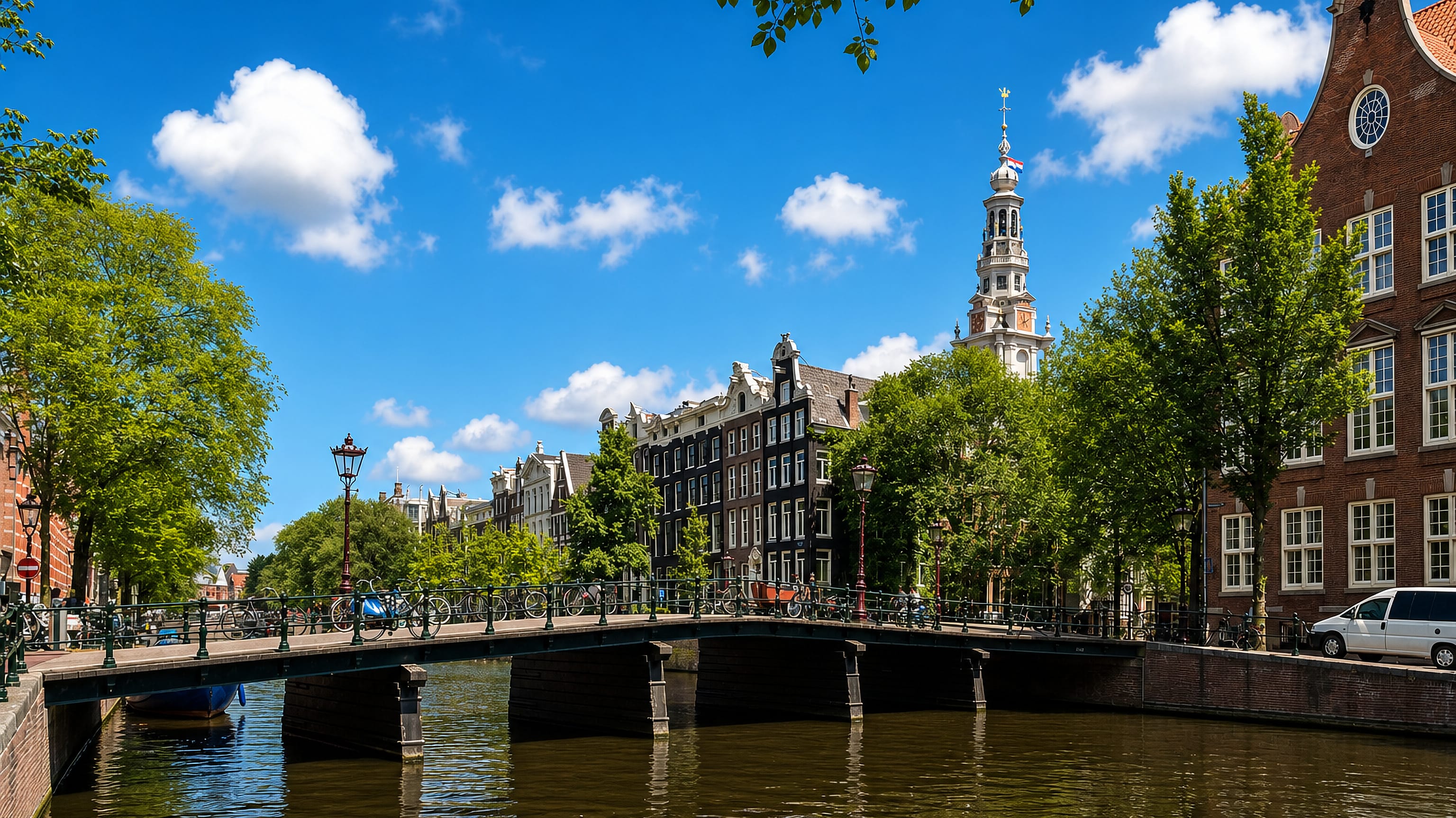 Canal bridge lined with bicycles below gabled canal houses and a church spire in Amsterdam's Grachtengordel.