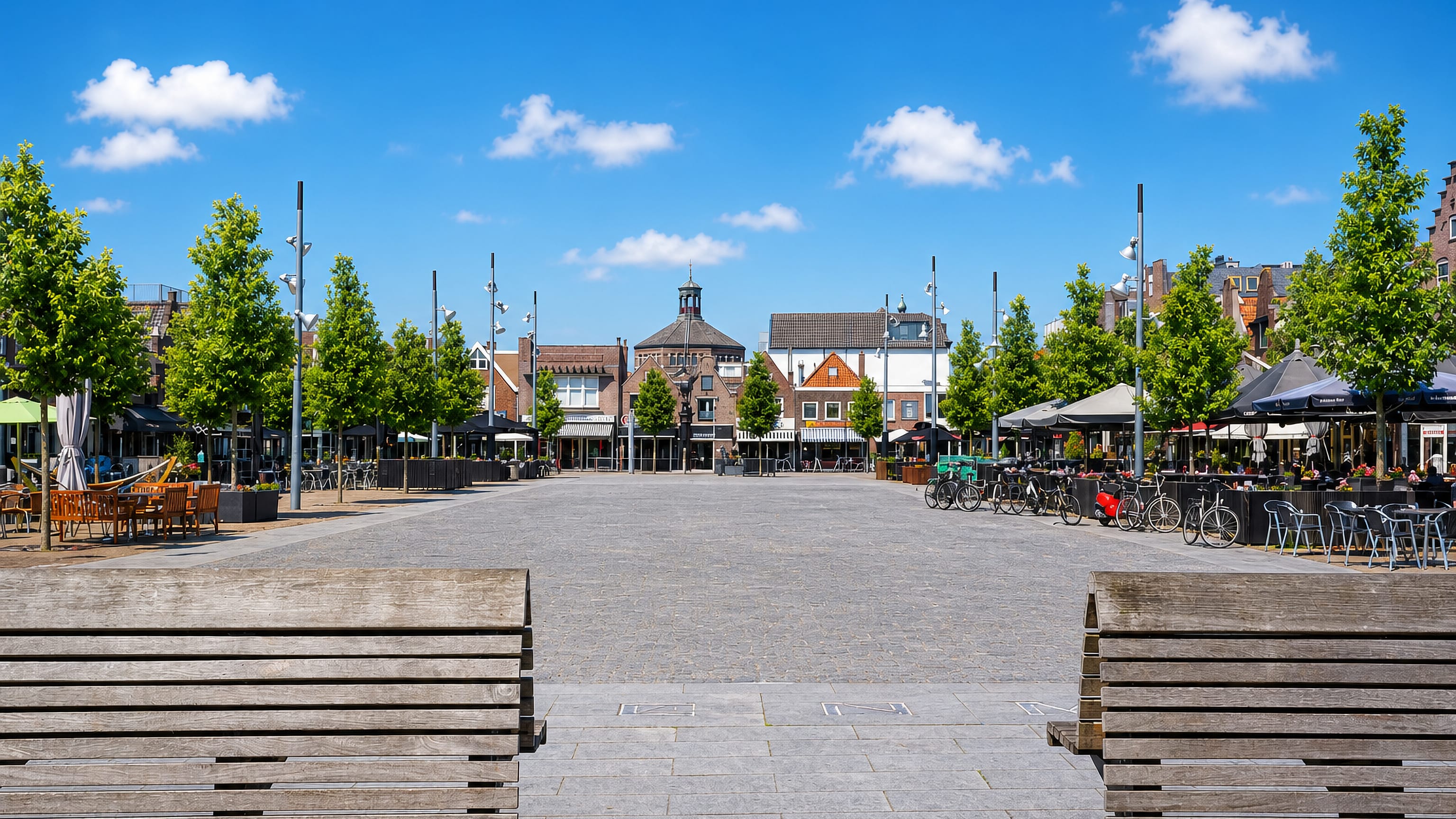 The wide paved market square of Purmerend lined with cafe terraces, trees and bicycles under a blue sky.