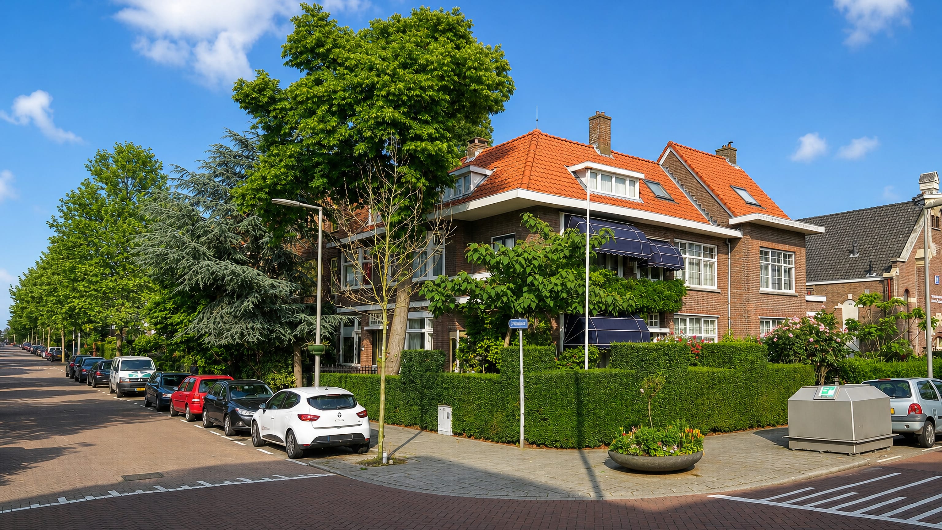Quiet brick street corner in Overschie, Rotterdam, with a 1930s villa, clipped hedges and parked cars.