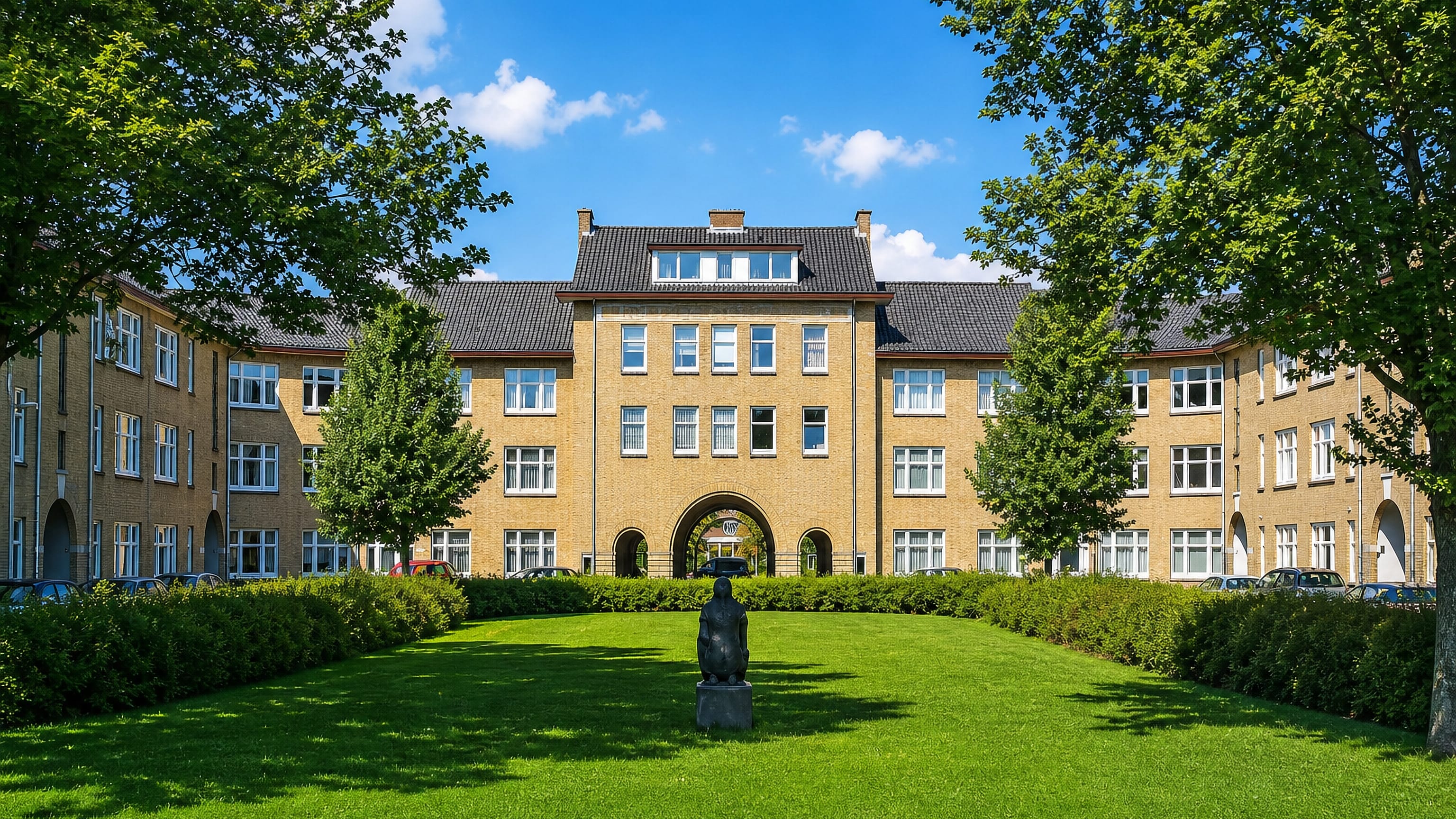Yellow-brick housing block with a central archway around a lawn and sculpture in Moerwijk, The Hague.