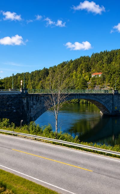Stone arch bridge over a calm river below forested hills near Åmli, Norway, beside a quiet roadside on a clear summer day.
