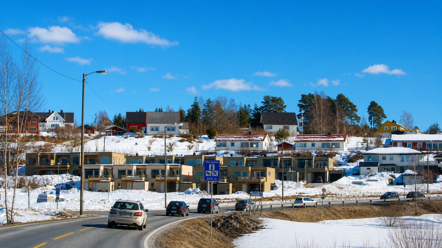 Winter roadside view of Birkeland, Norway, with modern townhouses on a snowy hillside and a place-name road sign.