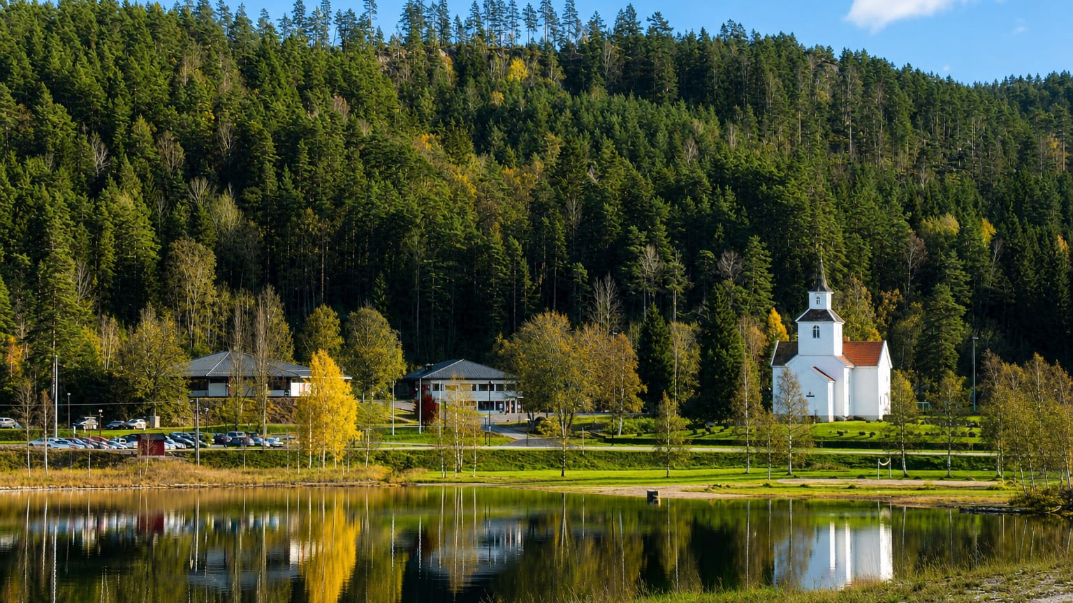 White lakeside church and timber houses below forested hills at Birketveit, Norway, mirrored in the calm lake