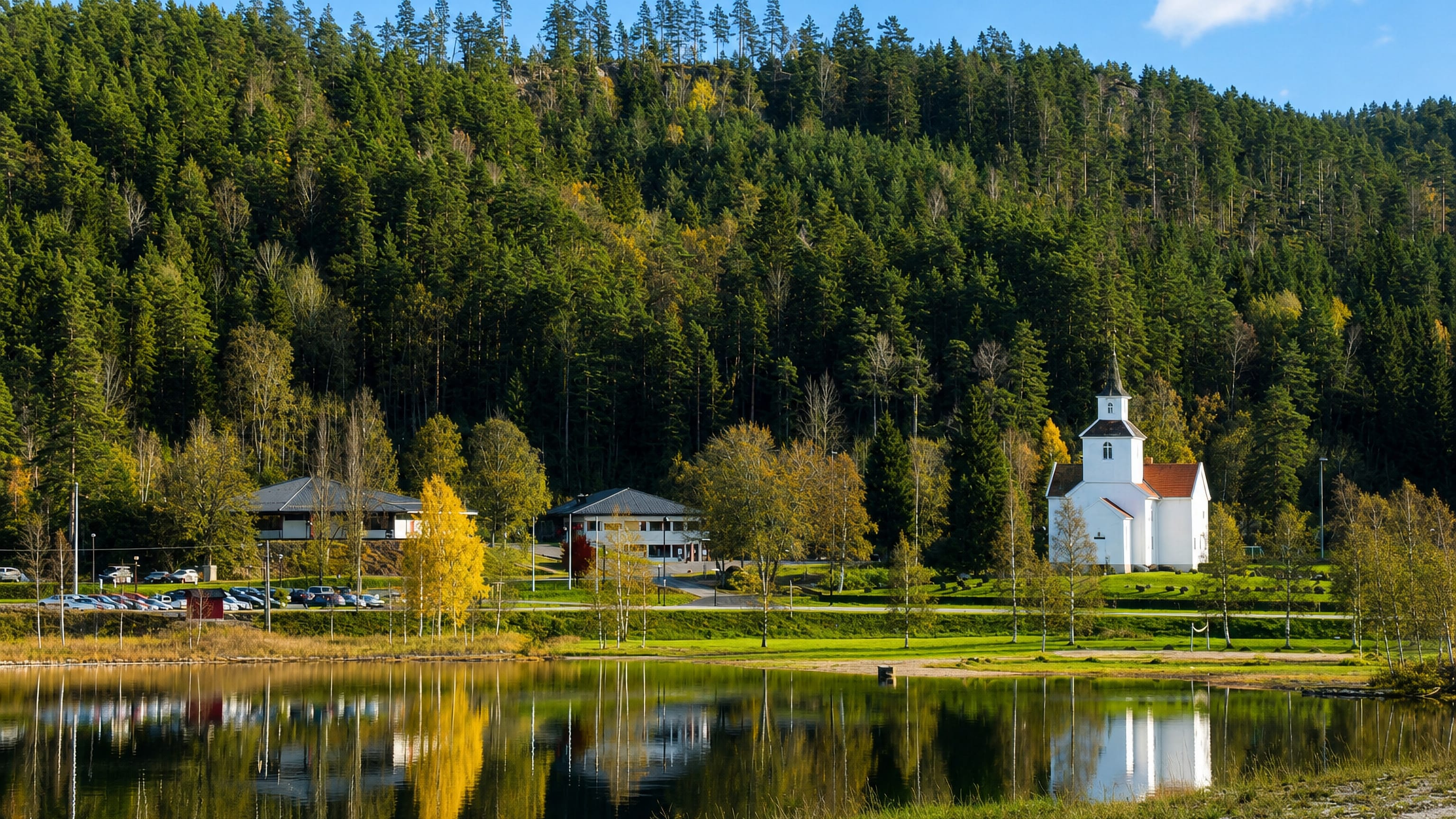 White lakeside church and timber houses below forested hills at Birketveit, Norway, mirrored in the calm lake