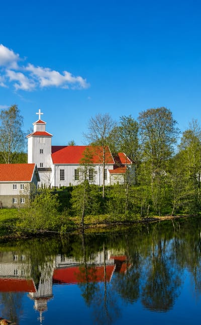 White wooden church with a red roof reflected in the calm Nidelva river amid forested hills at Blakstad, Norway
