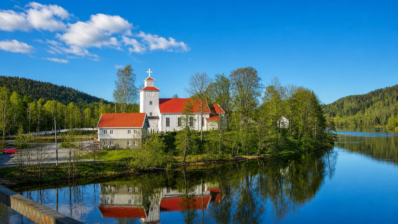White wooden church with a red roof reflected in the calm Nidelva river amid forested hills at Blakstad, Norway