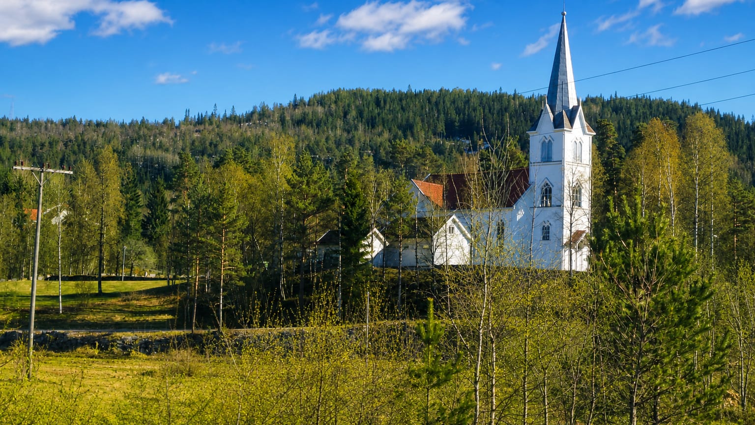 Evje's white wooden church with a tall steeple on a forested hillside above green meadows, Agder, Norway