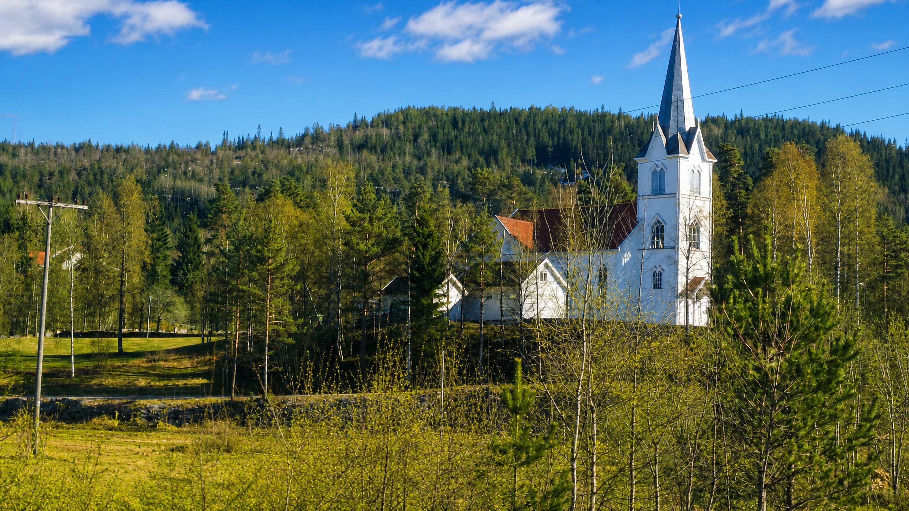 Evje's white wooden church with a tall steeple on a forested hillside above green meadows, Agder, Norway
