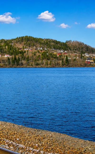 Lakeside view of Gjerstad, Norway, with scattered farms on forested hills and a railway line curving along the shore.