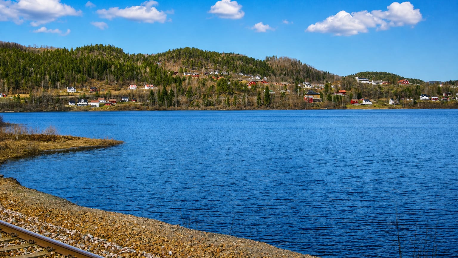 Lakeside view of Gjerstad, Norway, with scattered farms on forested hills and a railway line curving along the shore.