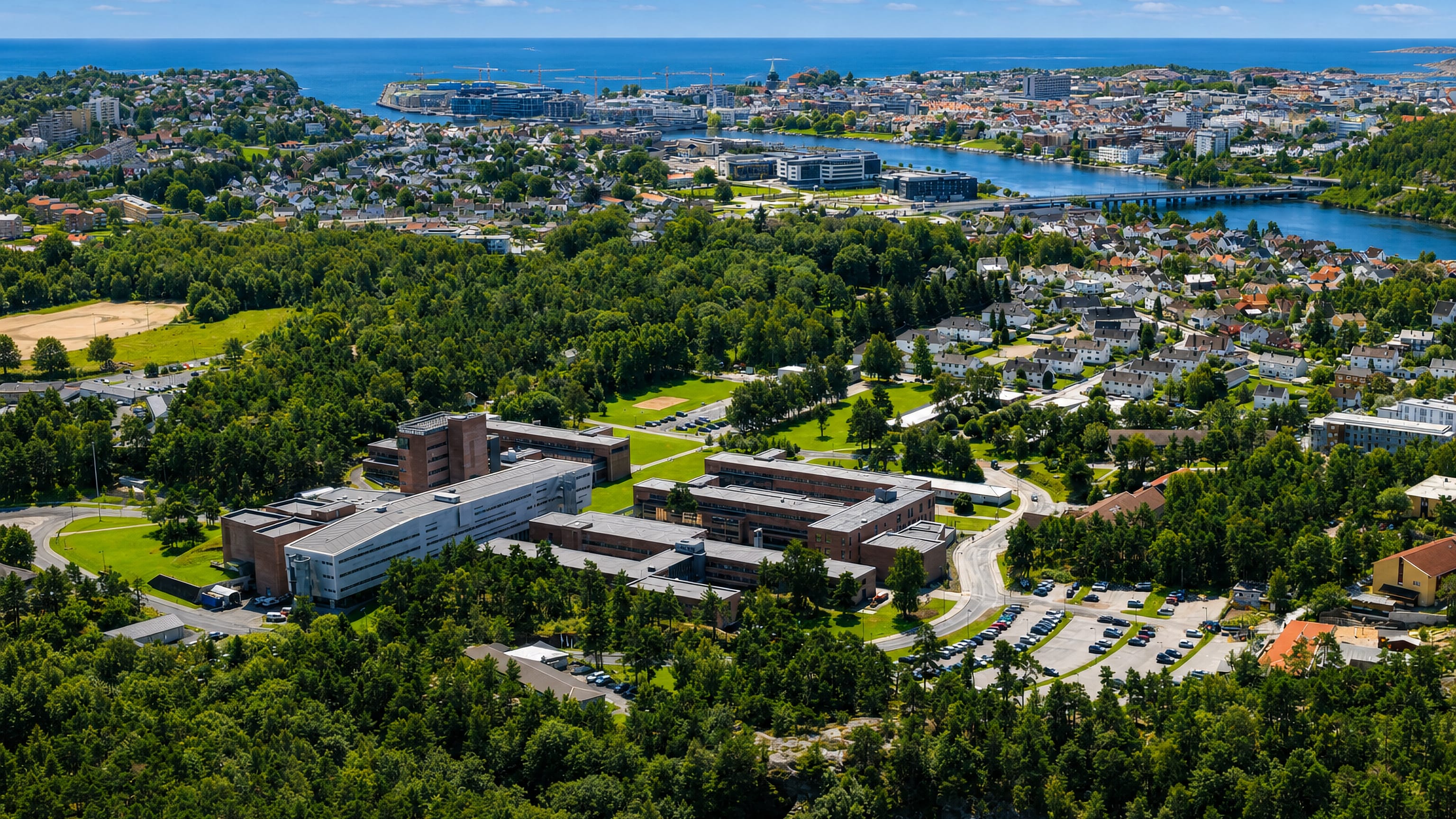 Aerial view of Lund in Kristiansand, campus buildings amid pine forest above the Otra river and the coastal centre.