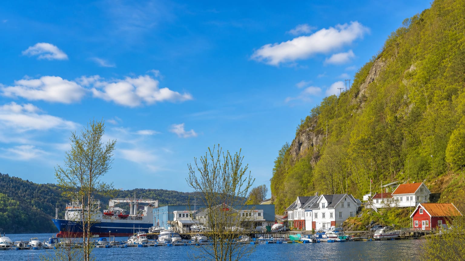 Waterside harbour with white and red houses, boats and a ship below a green hillside in Lyngdal, Norway.