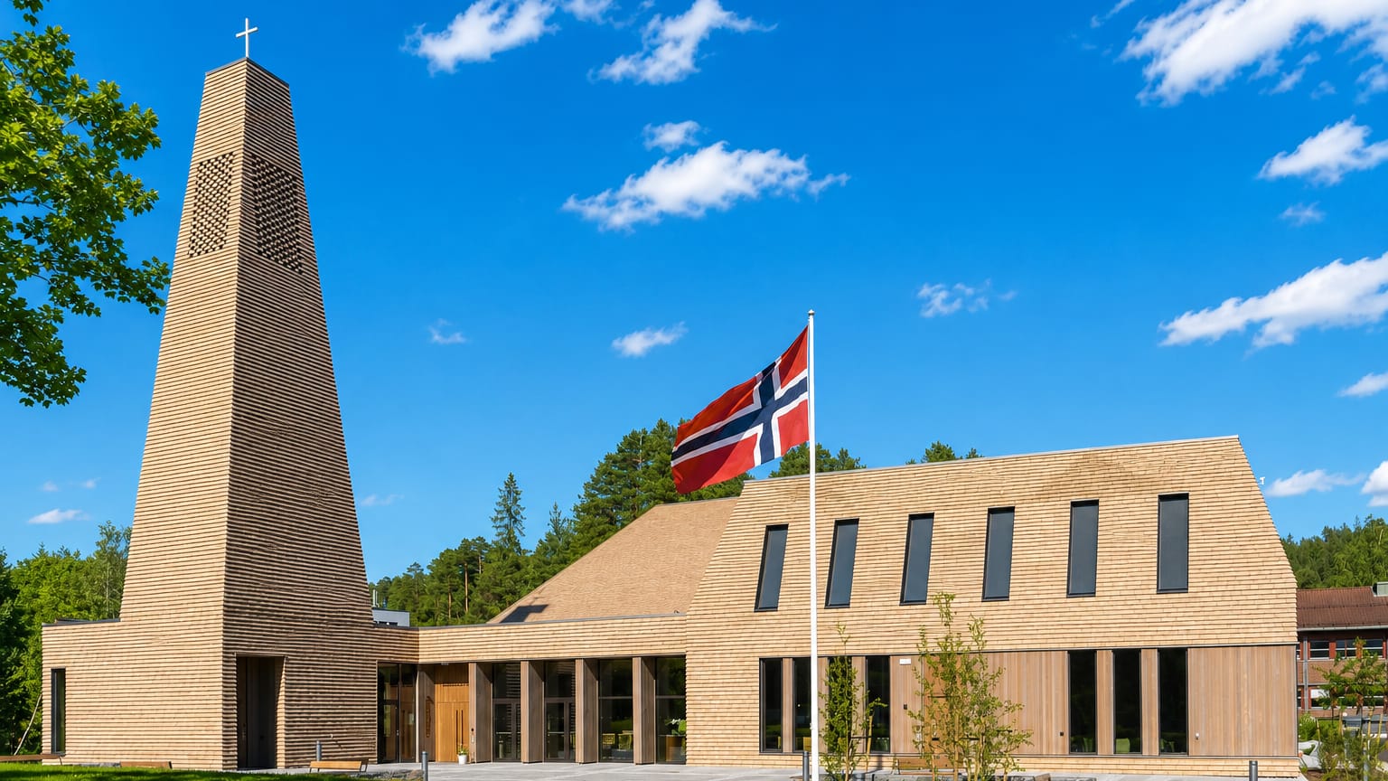 Modern timber-clad church with a tall pyramidal bell tower and Norwegian flag in Vennesla