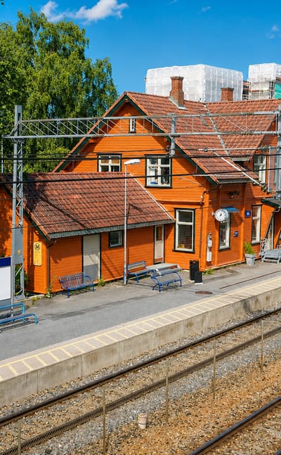 The orange wooden Ås railway station beside the train tracks and platform under a blue summer sky in Norway.