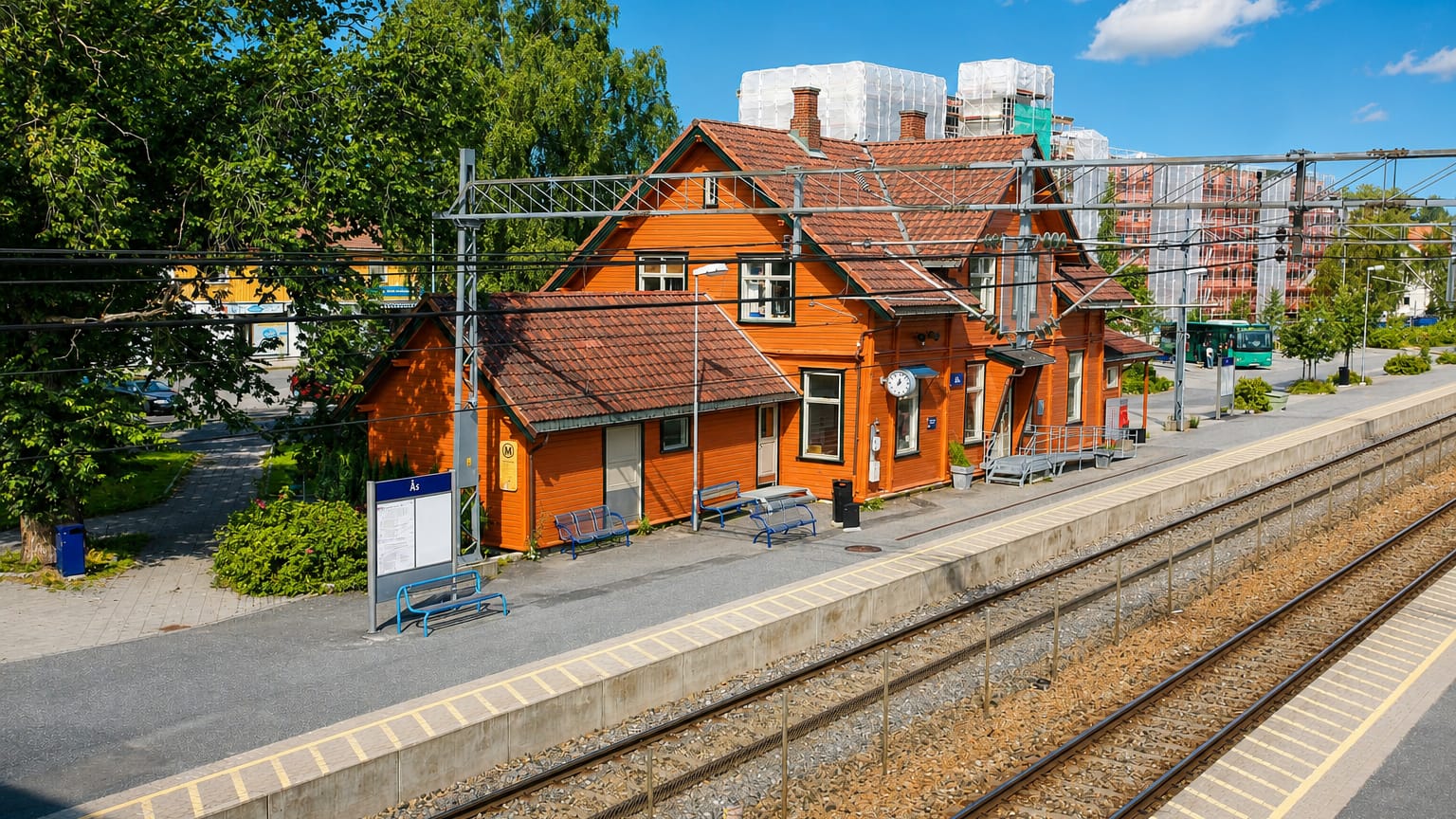 The orange wooden Ås railway station beside the train tracks and platform under a blue summer sky in Norway.