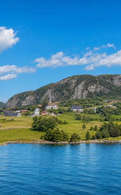 Blue fjord water fronting green meadows, scattered houses and a forested rocky hill at Asker, Norway.
