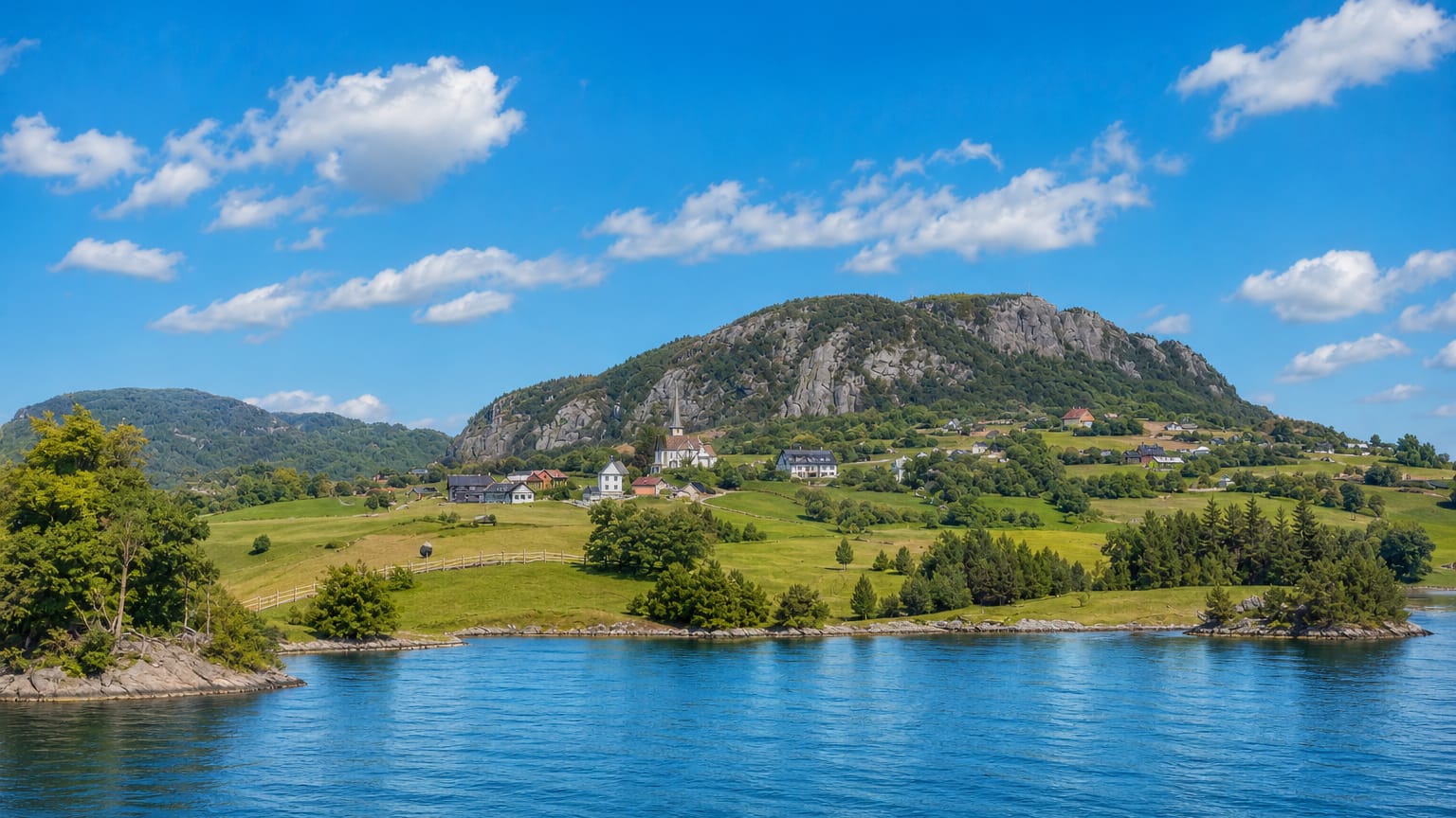 Blue fjord water fronting green meadows, scattered houses and a forested rocky hill at Asker, Norway.