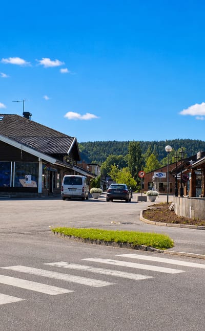 The wood-clad village centre of Bjørkelangen with a pedestrian crossing and forested hills behind.