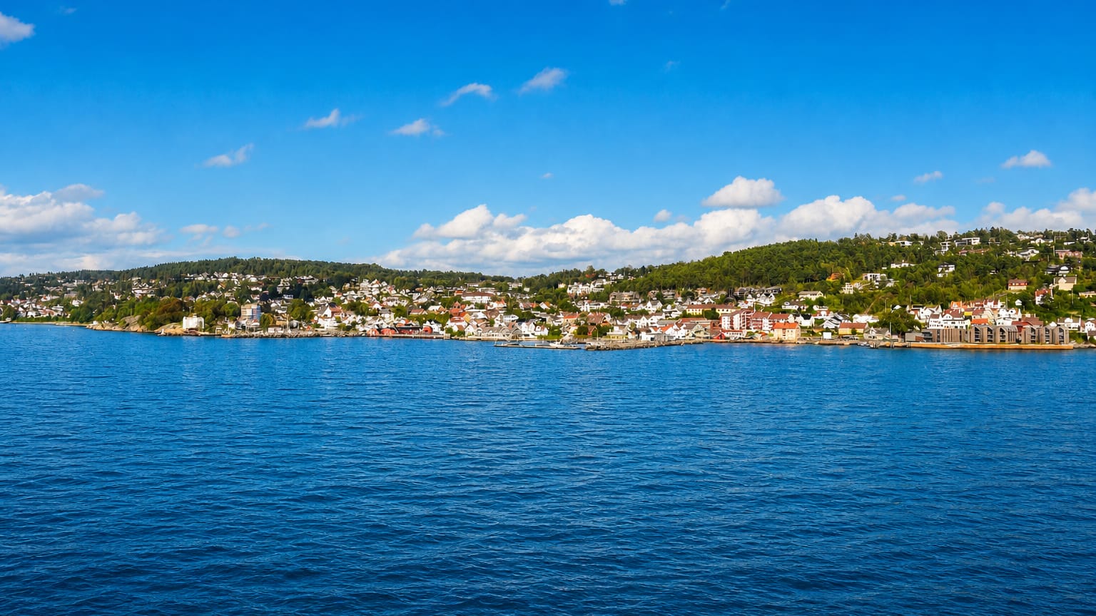 Distant view of Drobak, Norway, along the wooded Oslofjord shore across a broad expanse of blue water