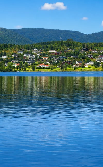 Hurdal's white houses scattered along the forested far shore of Lake Hurdalssjøen under a clear blue summer sky