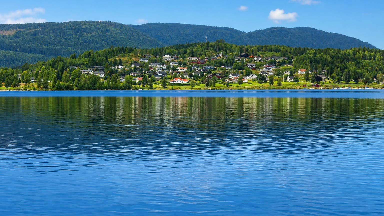 Hurdal's white houses scattered along the forested far shore of Lake Hurdalssjøen under a clear blue summer sky
