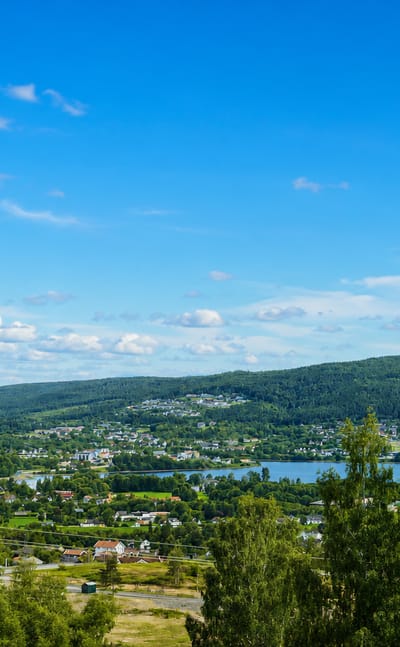 Elevated daytime view over Jevnaker, Norway, with the town beside Randsfjorden lake amid forested hills.