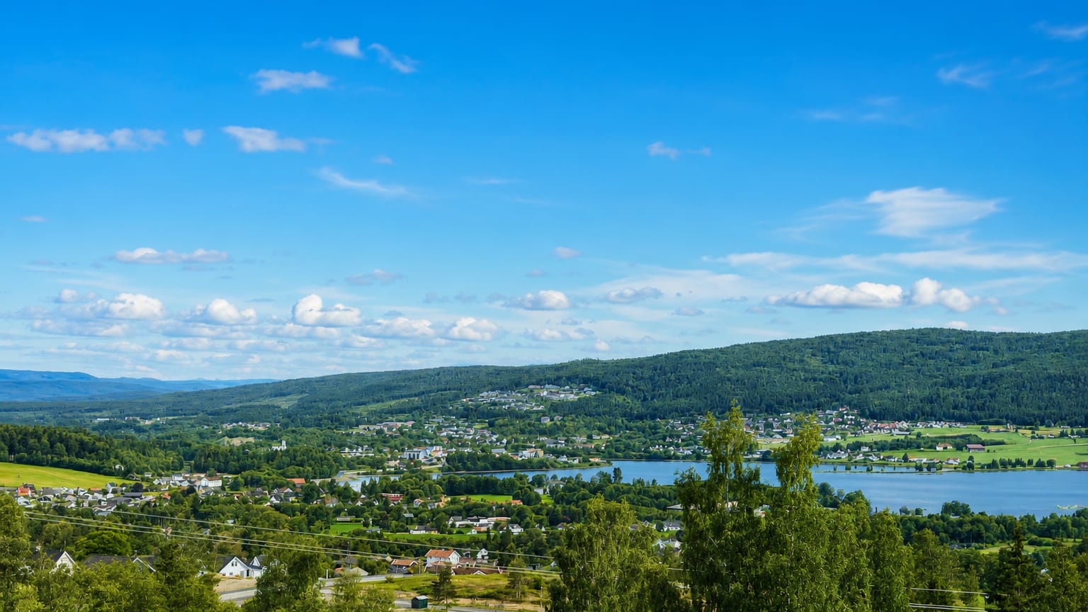 Elevated daytime view over Jevnaker, Norway, with the town beside Randsfjorden lake amid forested hills.