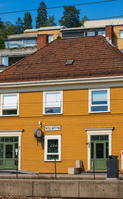 The yellow wooden Kolbotn railway station with its name sign, backed by tiered modern apartments on a wooded hillside.
