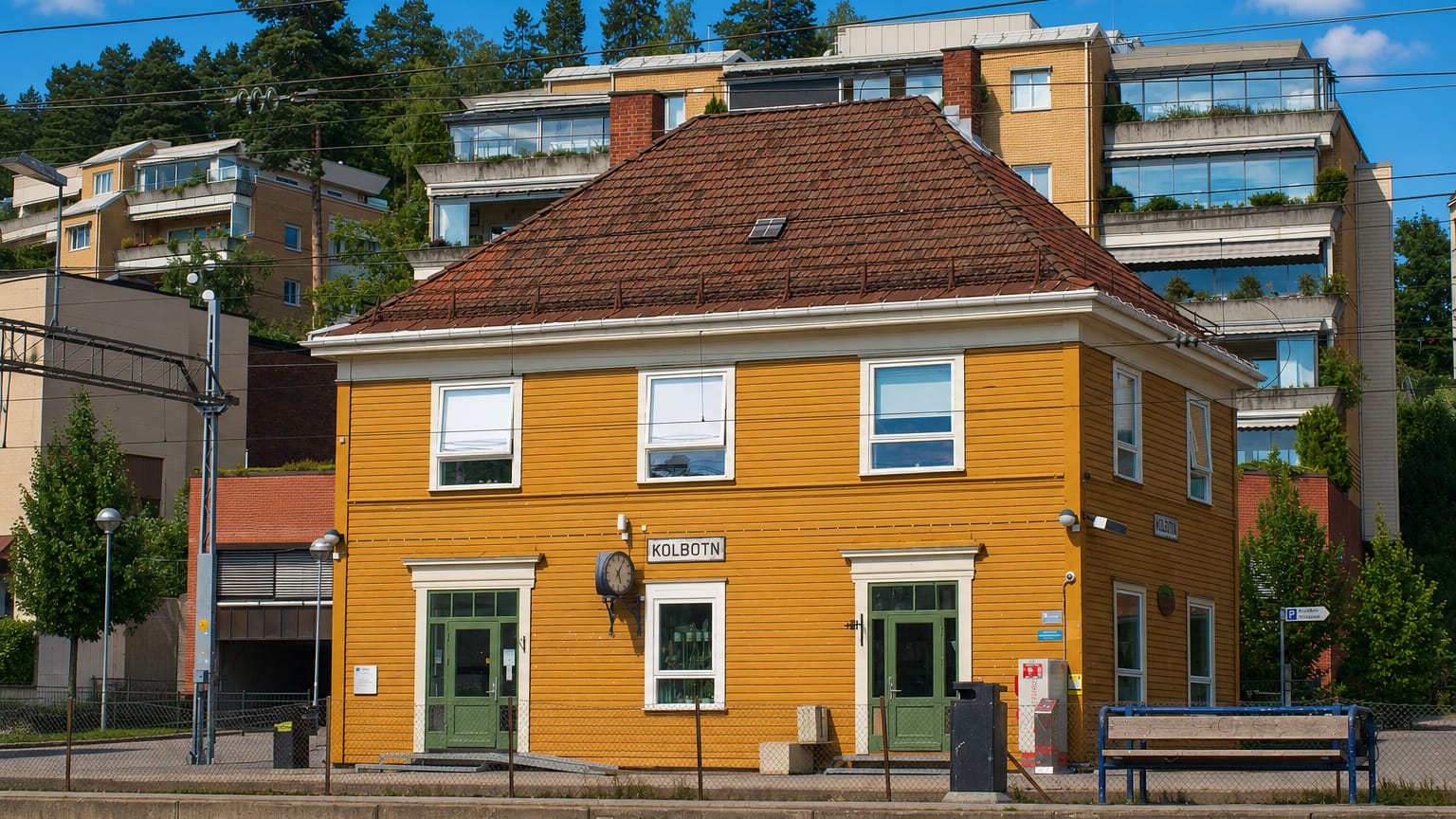 The yellow wooden Kolbotn railway station with its name sign, backed by tiered modern apartments on a wooded hillside.