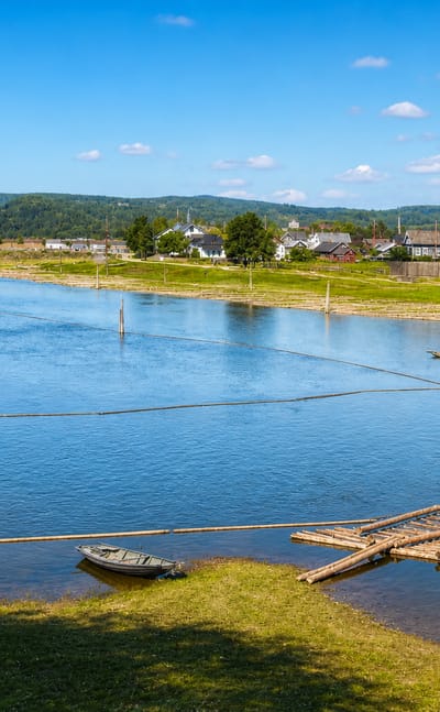 Wide daytime view of Lillestrøm, Norway: a river with a steel truss bridge, timber rafts and a town below forested hills.