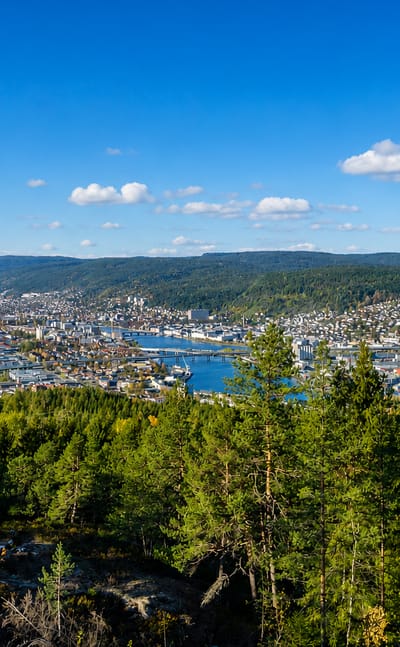 Aerial view of Drammen, Norway, spread across a river valley below forested hills under a blue sky