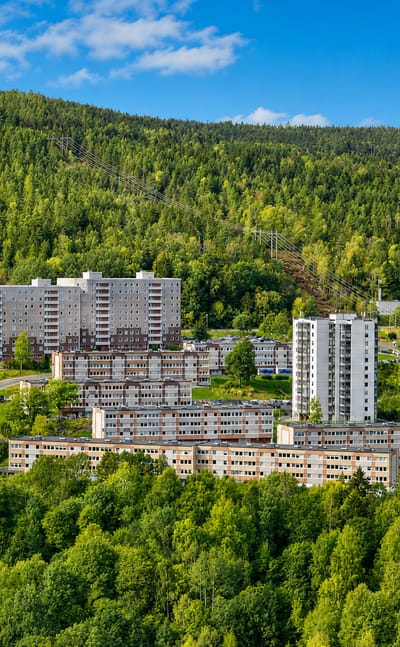 Concrete apartment tower blocks of the Fjell estate set into a forested hillside above Drammen under a clear blue sky.