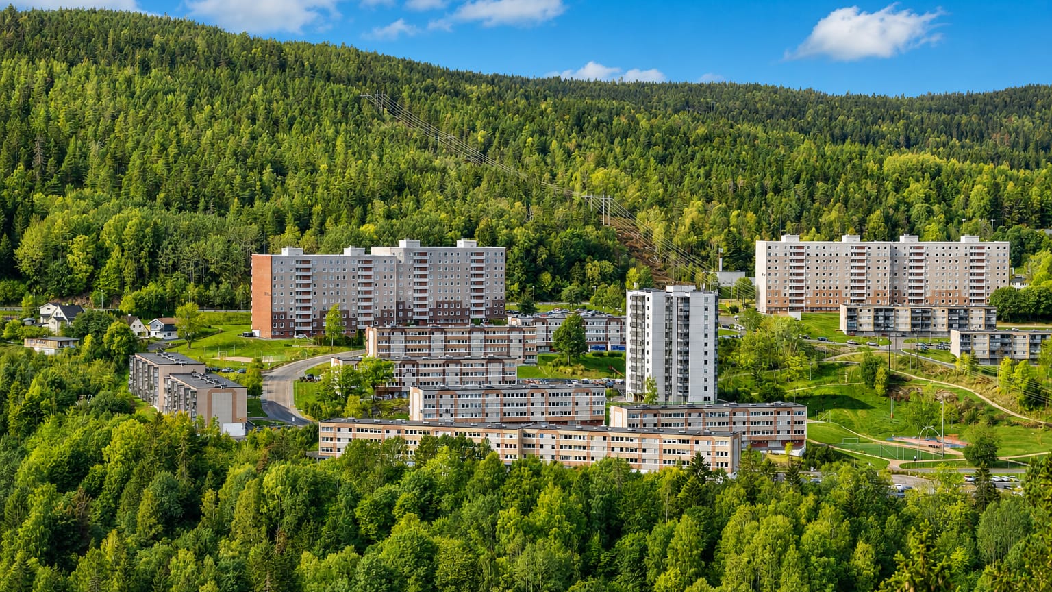Concrete apartment tower blocks of the Fjell estate set into a forested hillside above Drammen under a clear blue sky.