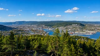 Aerial view of Drammen, Norway, spread across a river valley below forested hills under a blue sky