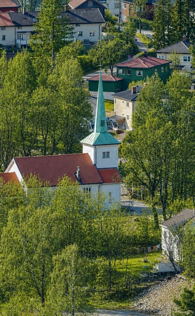 Strømsgodset Church with white walls and a green spire among lush trees and hillside homes in Drammen, Norway
