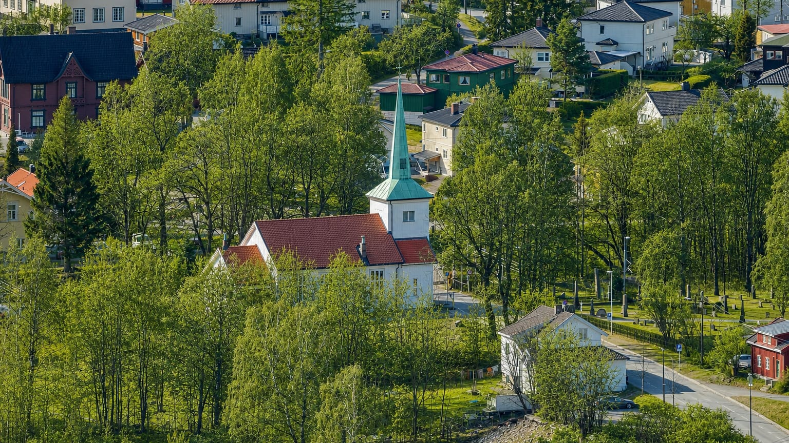 Strømsgodset Church with white walls and a green spire among lush trees and hillside homes in Drammen, Norway