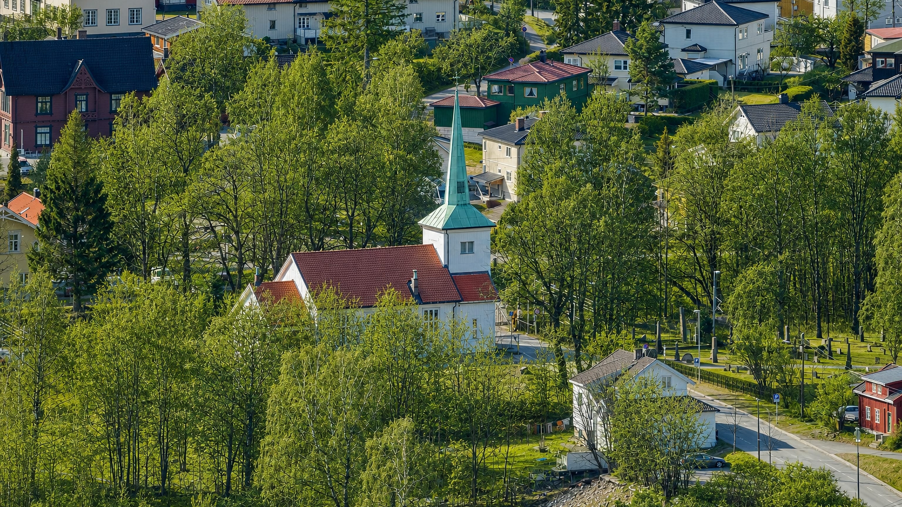 Strømsgodset Church with white walls and a green spire among lush trees and hillside homes in Drammen, Norway