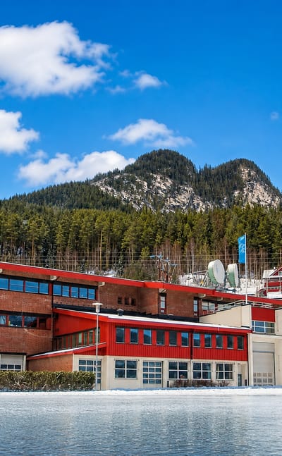 Red-clad modern buildings beside a snowy frozen lake below a forested hill in Gol, Norway, under a bright blue winter sky.