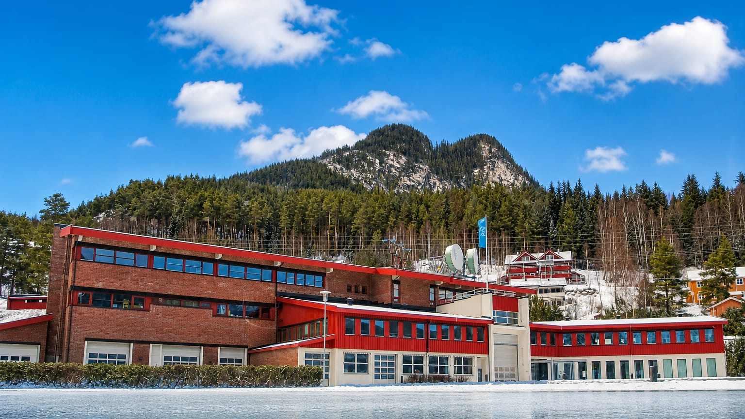 Red-clad modern buildings beside a snowy frozen lake below a forested hill in Gol, Norway, under a bright blue winter sky.