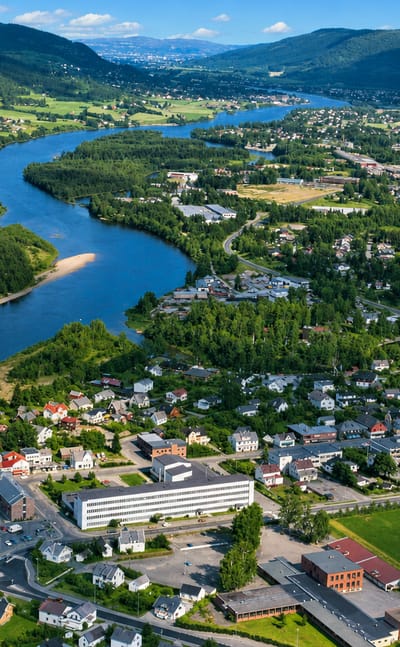 Aerial view of Hokksund, Norway, a riverside town along the Drammenselva winding through a green valley framed by forested hills.