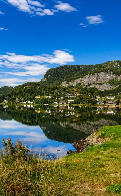 Calm lake mirroring forested mountains with a small lakeside village near Hol, Norway, under a clear blue summer sky.