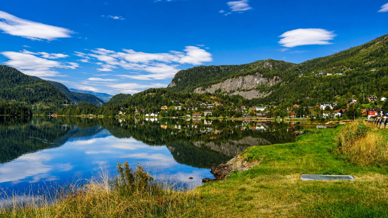 Calm lake mirroring forested mountains with a small lakeside village near Hol, Norway, under a clear blue summer sky.