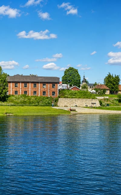 Hønefoss riverbank in Norway, with old brick and yellow industrial-era buildings, a grassy bank and a small church spire among the trees.