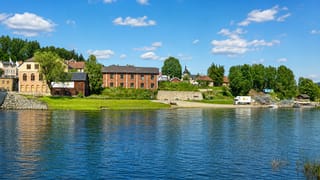 Hønefoss riverbank in Norway, with old brick and yellow industrial-era buildings, a grassy bank and a small church spire among the trees.