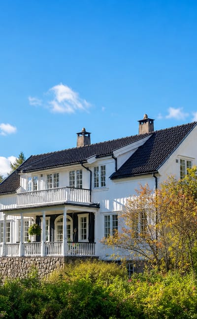 A white wooden villa with a balcony stands among autumn-tinged trees in Lampeland, Norway.