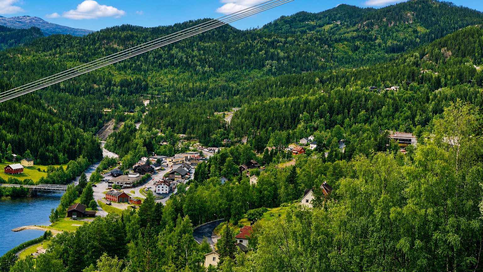 Aerial view of Rødberg, Norway, a small village clustered in a forested river valley framed by green mountains under a blue sky.
