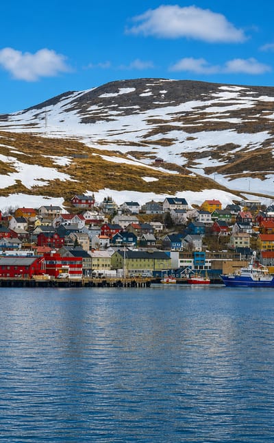 Honningsvag's colorful harbor houses and fishing boats below a snow-patched Arctic hill in Finnmark, Norway