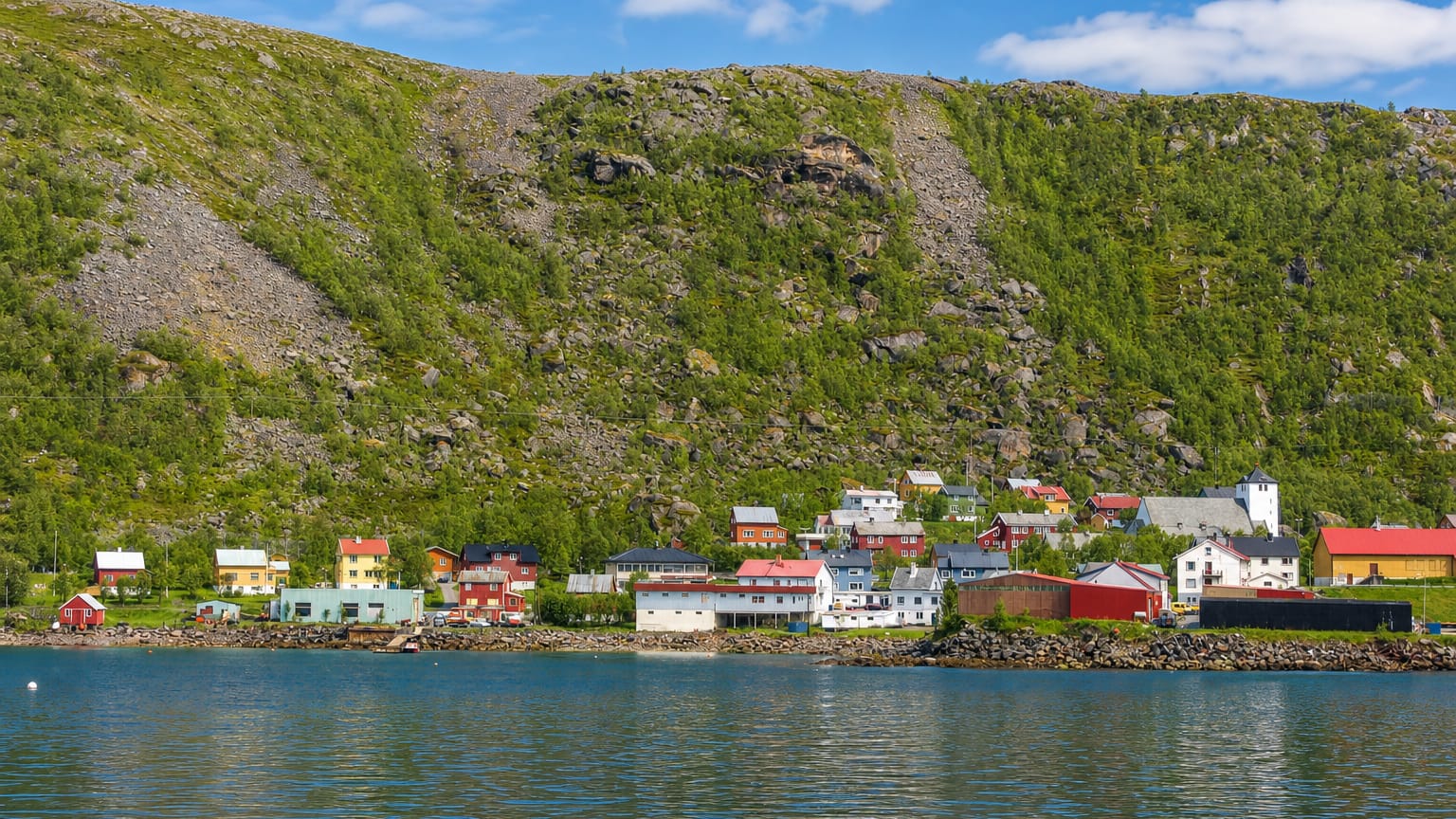 Øksfjord, Norway, its waterfront houses clustered beneath a steep green mountain along the fjord.